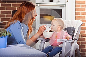 Beautiful mother in blue dress feeding with spoon her cute baby girl witch sits in high chair.