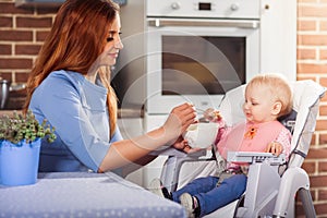Beautiful mother in blue dress feeding with spoon her cute baby girl witch sits in high chair.