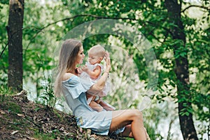 Beautiful mother in blue dress sitting on the ground in the summer forest on the hill and holding her baby on the hands