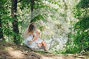 Beautiful mother in blue dress sitting on the ground in the summer forest and holding her baby on the hands