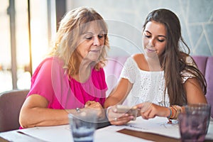 Beautiful mother and daugther sitting at restaurant using smartphone smiling