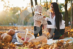 Beautiful mother is with her son in the autumn forest. Boy is holding orange leaf. Butternut squash and fruits on the ground