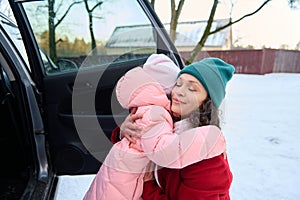 Beautiful mother hugging her daughter, standing together near car during a family trip on snow covered road, enjoying weekend on a