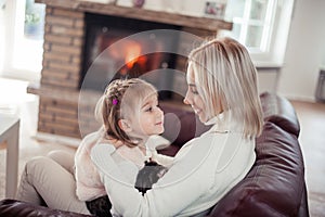 Beautiful mother, little daughter and a cat are sitting on the sofa near the fireplace. Family in the home interior. Cozy