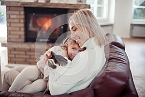 Beautiful mother, little daughter and a cat are sitting on the sofa near the fireplace. Family in the home interior. Cozy