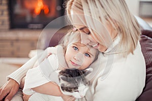 Beautiful mother, little daughter and a cat are sitting on the sofa near the fireplace. Family in the home interior. Cozy