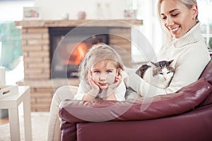 Beautiful mother, little daughter and a cat are sitting on the sofa near the fireplace. Family in the home interior. Cozy