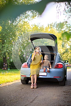 Beautiful mother and little son travelers sit in trunk of car on road and blow soap bubbles. Photo about family travel