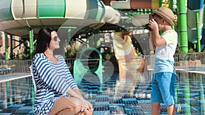 Beautiful mother posing at aquapark water slide background during cute son taking photo using camera