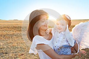 Beautiful mother with son walking in field. Boy in a pilot hat and angel wings sitting on hands of his mother