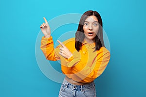 Beautiful multiethnicity woman in orange trendy shirt happily surprised expression pointing up against blue background