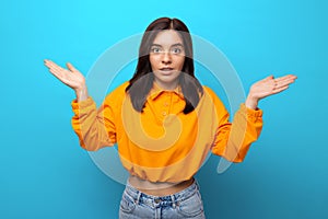 Beautiful multiethnicity woman in orange trendy shirt surprised expression against blue background