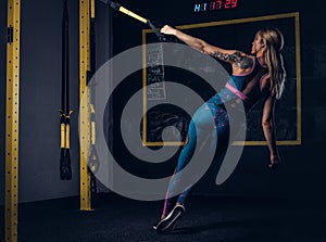 Beautiful muscular blonde woman in sportswear with a tattoo on her arm doing exercise with TRX system at gym.