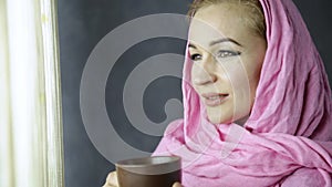 Beautiful muslim arabian woman in pink hijab drinks cofee in front of window