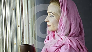 Beautiful muslim arabian woman in pink hijab drinks cofee in front of window
