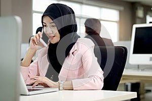 Beautiful Muslim Asian woman officer with hijab, talking with customer by mobile phone while working on laptop computer at office