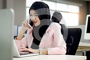 Beautiful Muslim Asian woman officer with hijab, talking with customer by mobile phone while working on laptop computer at office