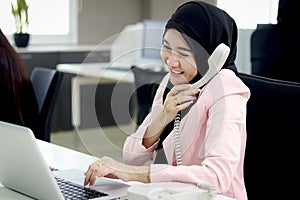 Beautiful Muslim Asian woman officer with hijab, talking on landline phone with customer while working on laptop computer at