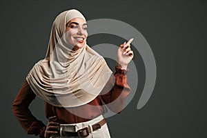 Beautiful muslim woman in beige hijab pointing finger at space on gray background. Copy space