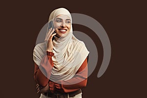 Beautiful muslim woman dressed in beige hijab holding phone smiling and talking on brown background. Copy space