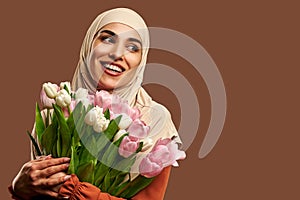 Beautiful Muslim woman dressed in a beige hijab smiles and looks away and holds a large bouquet of tulips on a brown background.