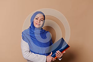 Beautiful Muslim woman in hijab, female confident educator teacher holds books and looks at camera, on beige background