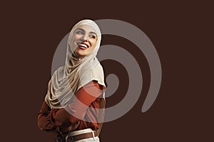 Beautiful muslim woman wearing beige hijab, orange blouse and beige pants smiling and looking back on brown background. Copy space