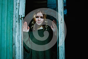 A beautiful mysterious blonde woman in a vintage sweater stands outside the window in the darkness of an abandoned old creepy