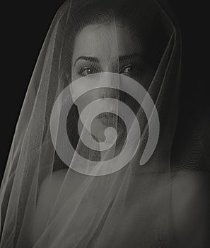 Beautiful mysterious woman face in darkness covered white veil on dramatic shadow black background. Closeup emotion studio