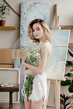 beautiful naked young woman holding white flowers and looking at camera in art studio