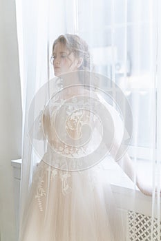 Beautiful natural redhead girl bride, with nude makeup, wearing a white dress, stands at the window behind a transparent curtain