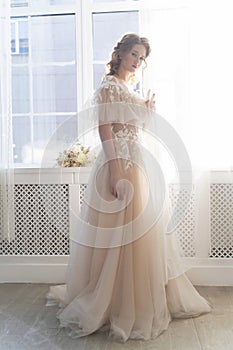 Beautiful natural redhead girl bride, with nude makeup, wearing a white dress, stands at the window next to a transparent curtain