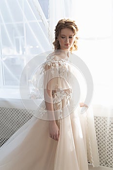 Beautiful natural redhead girl bride, with nude makeup, wearing a white dress, stands at the window next to a transparent curtain