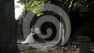 Beautiful newlyweds are sitting on an old bench in a green park. Action. The stylish groom embraces the lovely bride in