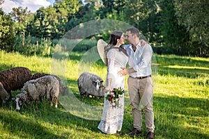 Beautiful newlyweds stand against the background of a fence with green foliage. The stylish bridegroom holds the