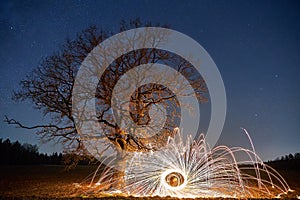 Beautiful night landscape with a lonely tree against the background of stars and a fire show. Alternative landscape