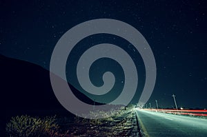 Beautiful night landscape of stars at sky and mountain silhouette near road with car trails. Road in the mountains under a starry