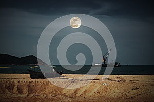 Beautiful night sky with bright full moon above cargo ship anchored in the sea. Traditional fishing boat parked on tropical beach