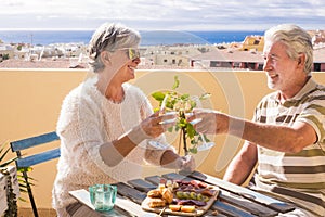 Beautiful old mature couple enjoy together clinking and roasting cocktails outdoor at home at the terrace with city and ocean view