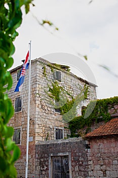 Beautiful old stone house in the village of Milna overgrown with green plants, croatian flag on a pole waving in front of it