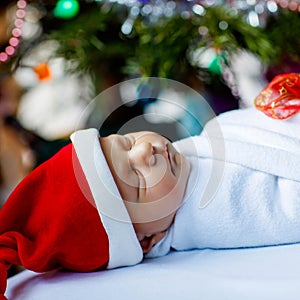 Beautiful one week old newborn baby wrapped in blanket near Christmas tree with colorful garland lights on background