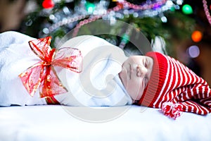 Beautiful one week old newborn baby wrapped in blanket near Christmas tree with colorful garland lights on background