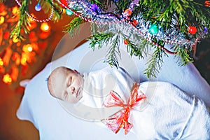 Beautiful one week old newborn baby wrapped in blanket near Christmas tree with colorful garland lights on background