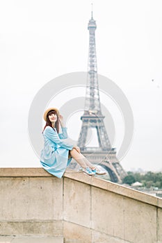 Beautiful parisian red haired girl in stylish hat and blue dress sitting at the stairs with the Eiffel tower background