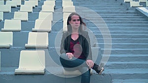 Beautiful and passionate girl in a biker jacket alone sits in an empty stadium. Girl`s hair develops in the wind.