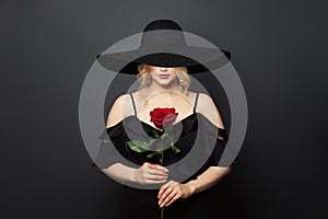 Beautiful passionate woman wearing wide black broad brim hat with red rose flower on black background