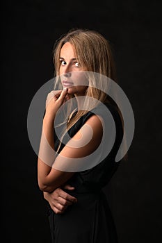 A beautiful pensive blonde girl stands in half a turn on a black background and looks into the camera