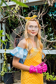 beautiful pensive blonde woman in apron and rubber gloves holding scissors and looking away