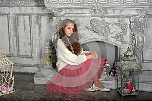 A beautiful pensive girl in a white sweater and pink skirt by the fireplace