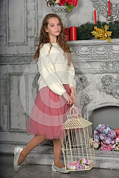 A beautiful pensive girl in a white sweater and pink skirt by the fireplace